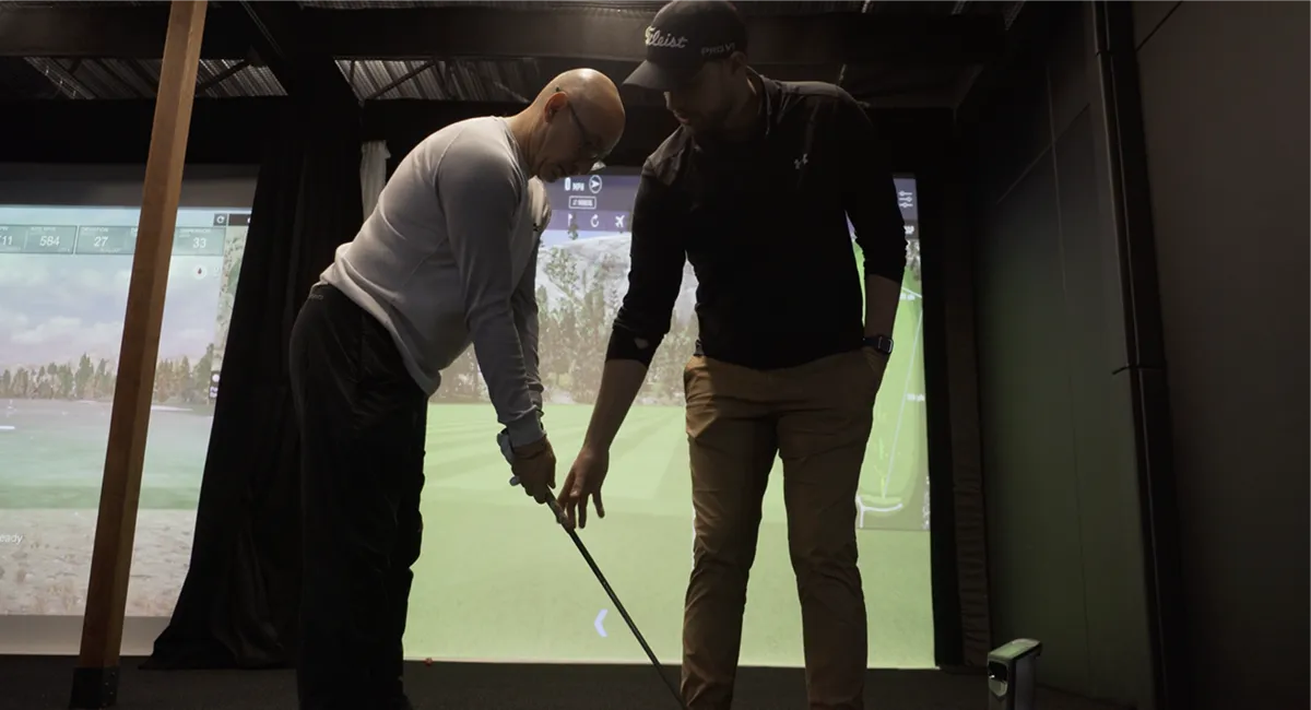 A golf instructor adjusting a student's grip and club position during a lesson in an indoor golf simulator.