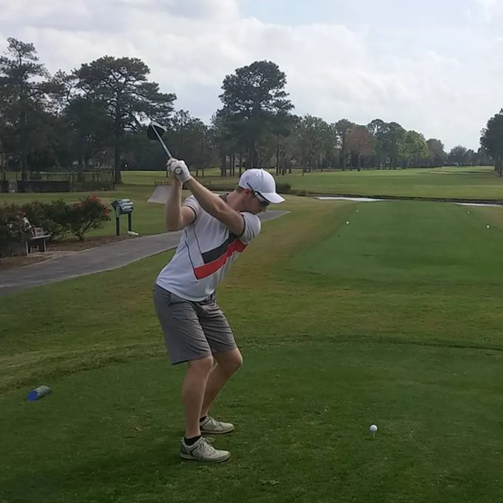 A young golfer in a white cap and grey shorts at the top of his backswing on a green fairway lined with trees.