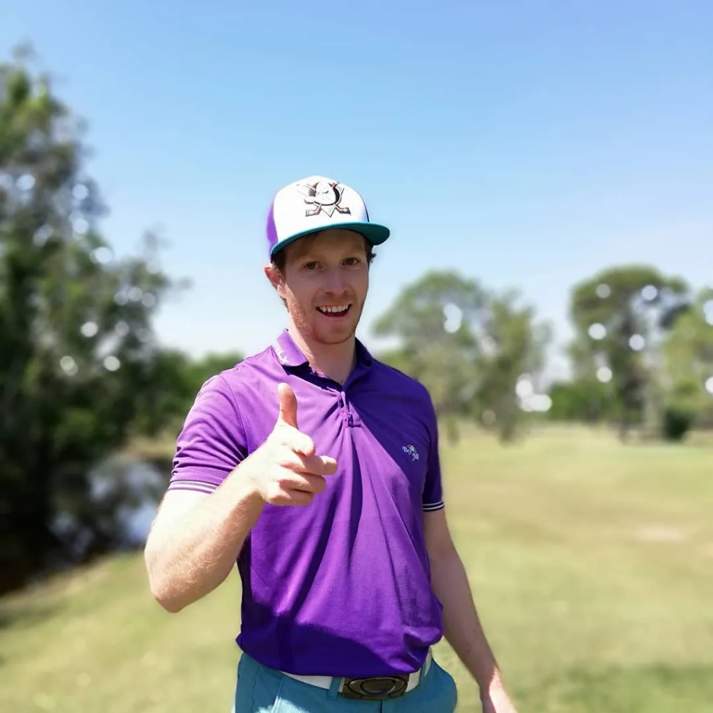 A smiling man in a purple polo and a white and teal trucker hat giving a thumbs-up on a sunny golf course.