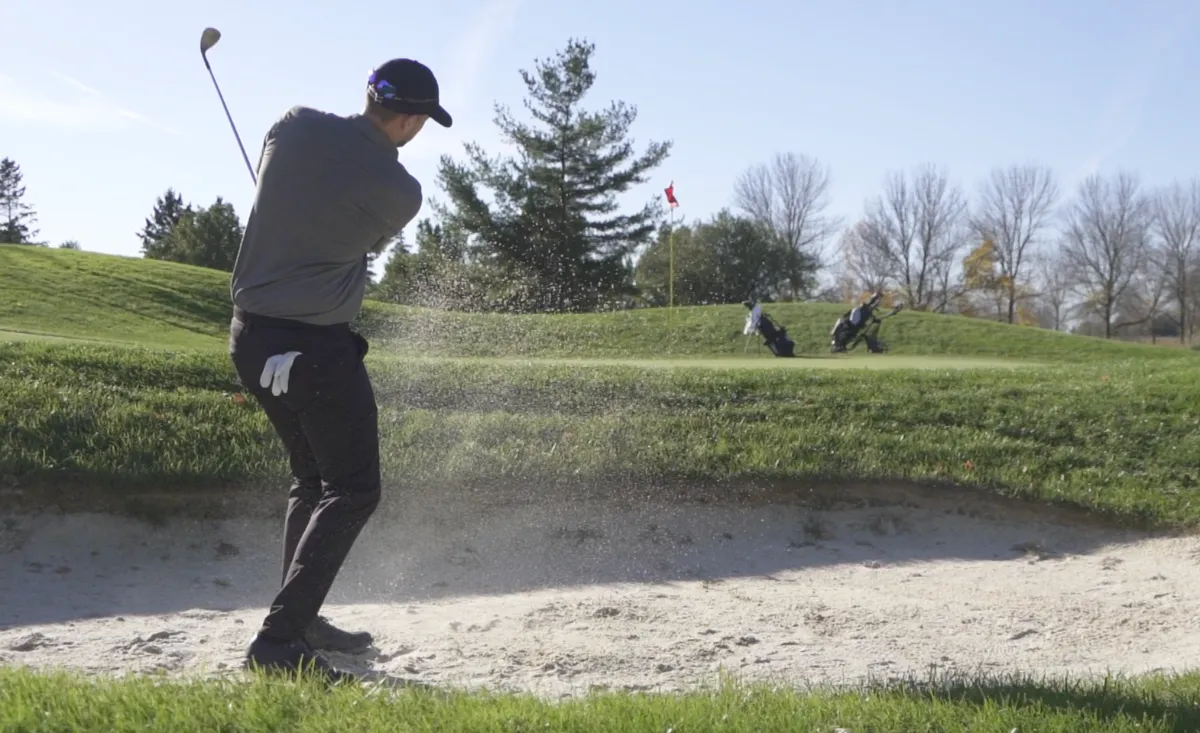 A golfer hitting a shot out of a sand bunker toward the green on a sunny day.