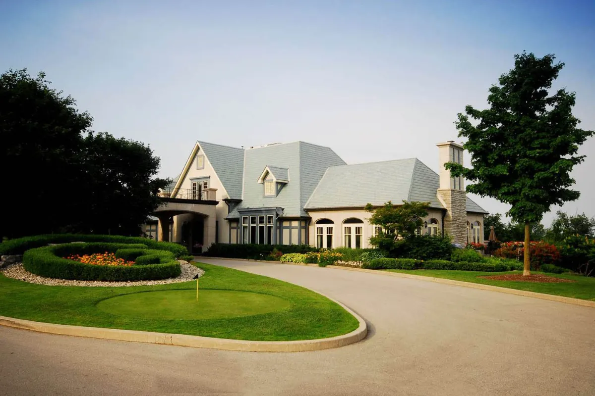A grand, multi-story golf clubhouse with light grey shingles and stone accents stands behind a manicured circular driveway and a small practice putting green.