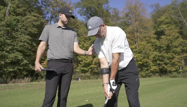 A golf instructor guiding a student's arm alignment and setup position during an outdoor lesson.