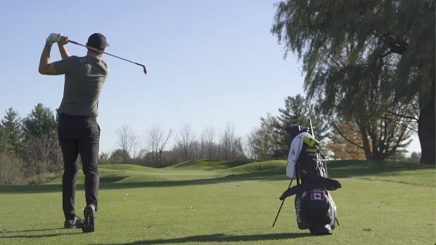A golfer completing a full follow-through swing on a sunlit course with a golf bag resting in the foreground.