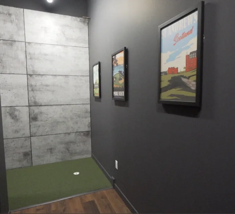 An indoor putting green located in a modern hallway with dark grey walls. Three framed travel-style posters are mounted on the wall, leading toward a small green turf area with a single hole and a concrete-textured accent wall.
