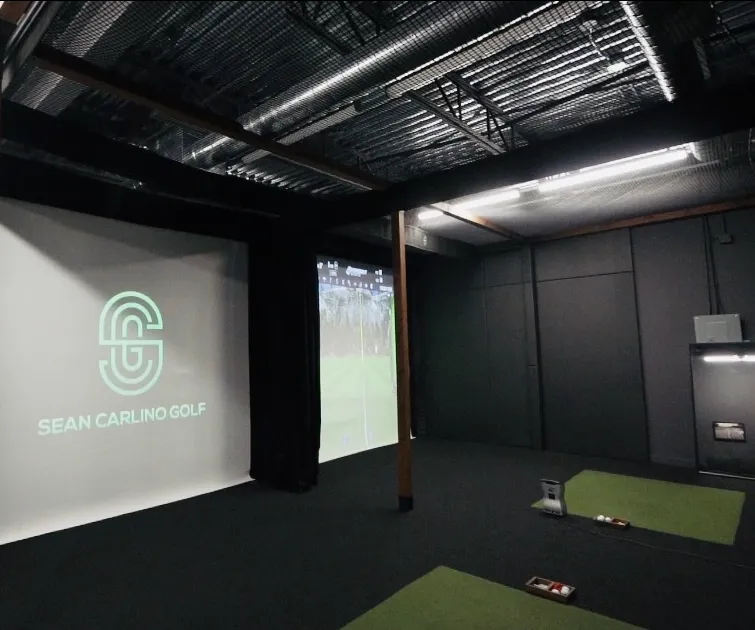 An industrial-style indoor golf facility featuring two hitting bays with green turf mats, black walls, and a large projection screen displaying the 'South Side Golf' logo.