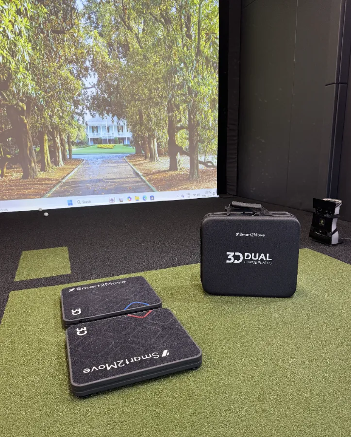 A high-angle shot of professional golf simulator equipment, including a launch monitor and two hitting mats, set up in front of a large digital projection screen showing a tree-lined course.