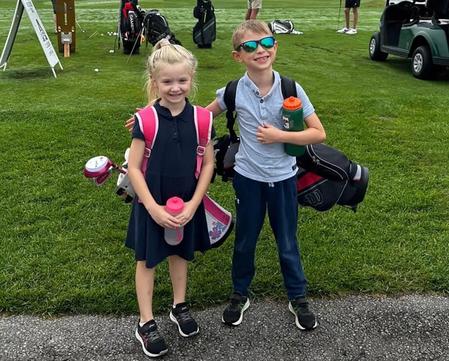 A young girl in a navy dress and a young boy in a light blue polo standing together on a grass golf course. Both are carrying golf bags and holding water bottles, with a golf cart and other equipment visible in the background.