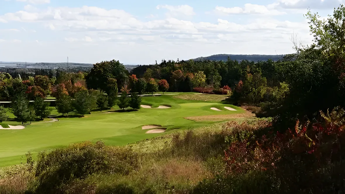 A panoramic view of a lush green golf course fairway with several sand bunkers, surrounded by dense forests and rolling hills under a bright, partly cloudy sky.