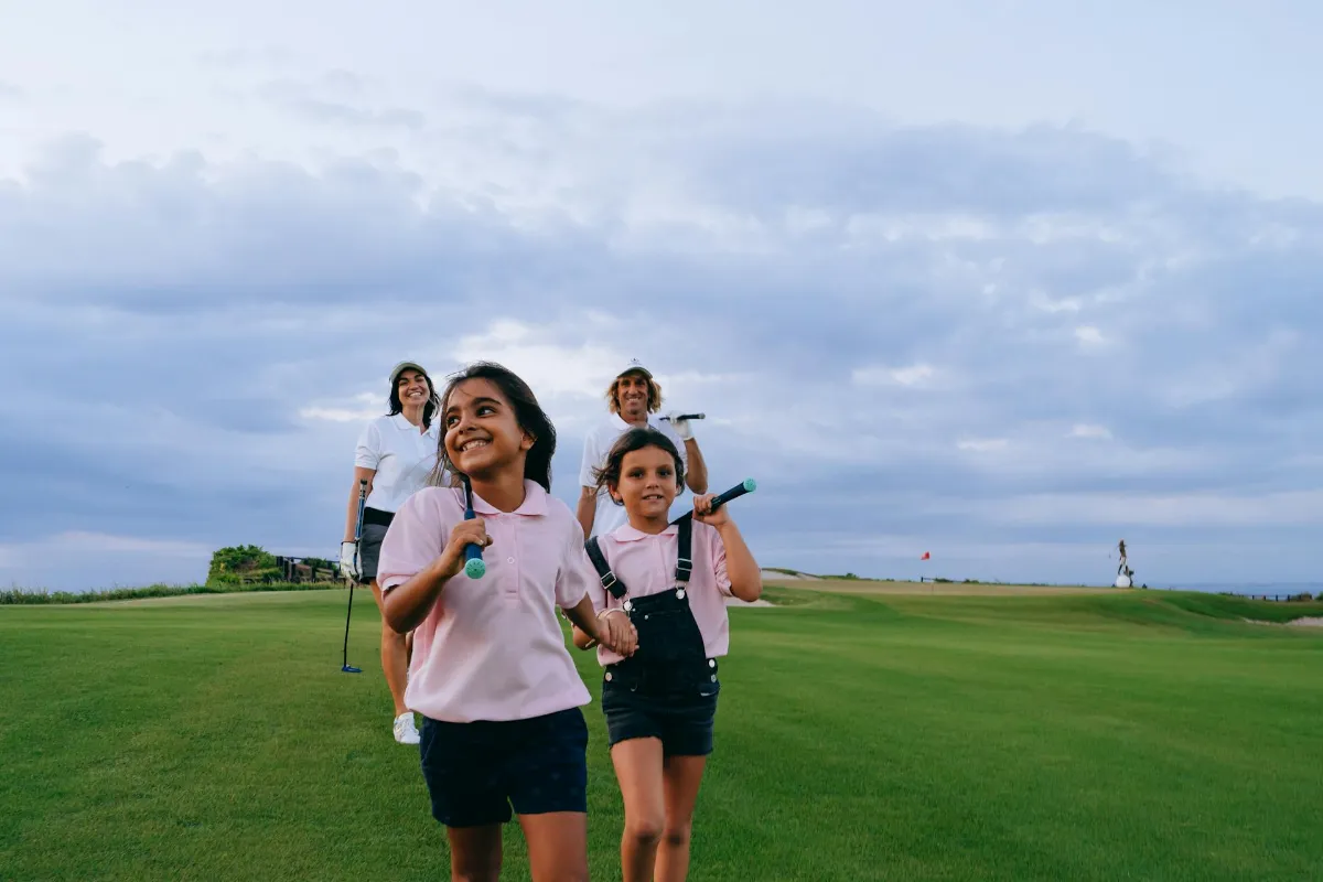 A smiling family of four walks across a lush golf course under an overcast sky, with two young girls in pink polo shirts leading the way while carrying golf clubs.