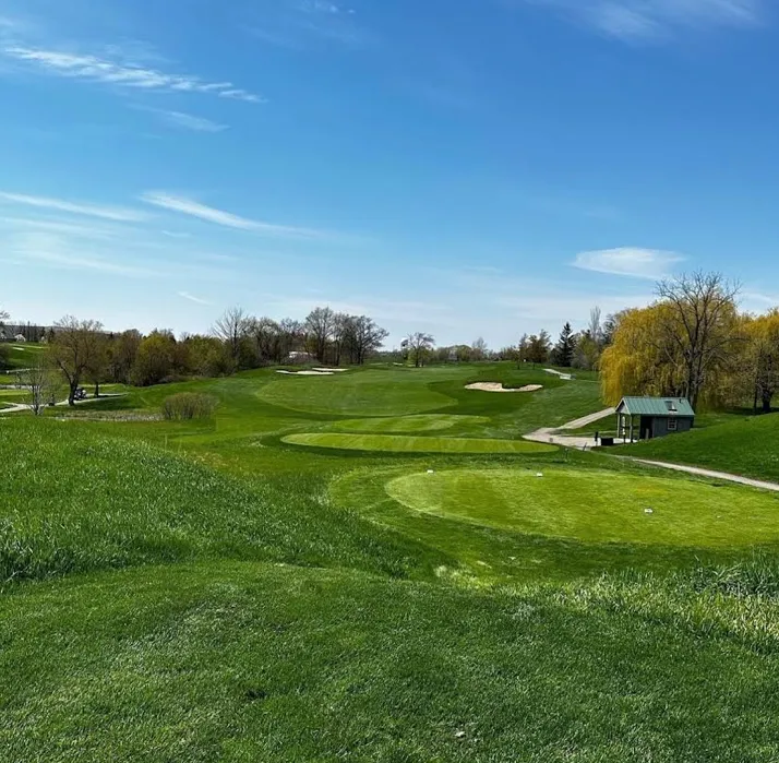 A wide-angle landscape shot of a lush, green golf course under a bright blue sky with wispy clouds, featuring rolling fairways, sand bunkers, and a small wooden structure nestled among trees on the right.