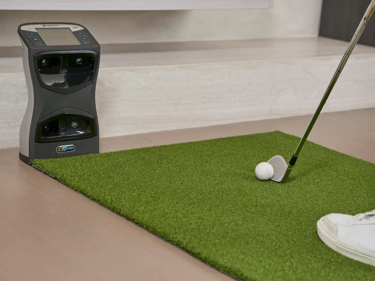 A launch monitor sits next to an indoor hitting mat where a golfer is about to strike a ball with a club.